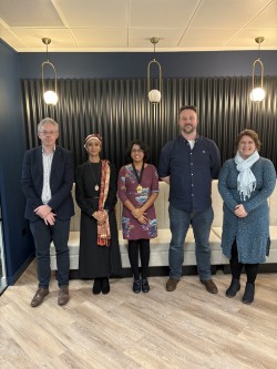 Image Description: A group of five people from Bristol Law Society Executive Council standing in front of a modern black-panelled wall with decorative lighting with the president and vice president wearing presidential gongs around their necks. A light-coloured bench is behind them on a wood-patterned floor.